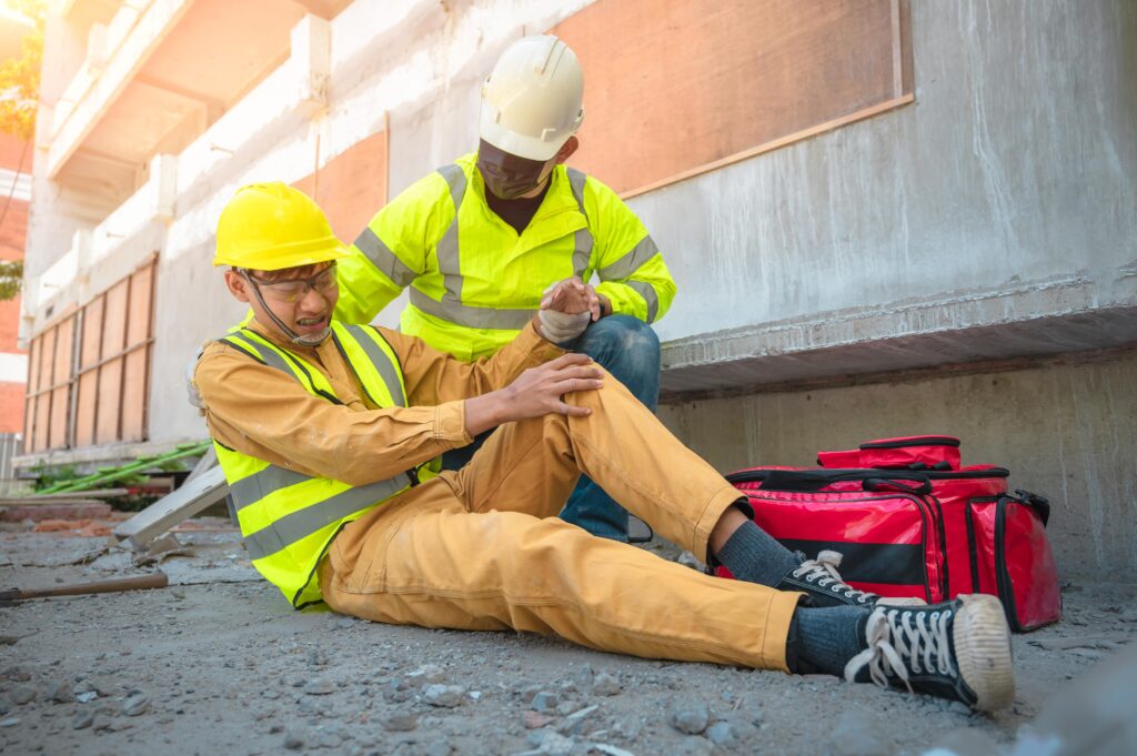 Dos trabajadores de la construcción con cascos de seguridad y chalecos reflectantes. Uno de ellos está sentado en el suelo con una expresión de dolor en la pierna, mientras el otro lo asiste y le sujeta el brazo. Cerca hay una bolsa roja de primeros auxilios.