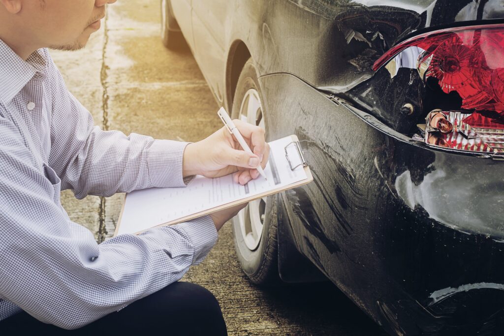 Un hombre con una camisa de cuadros y pantalones oscuros está agachado junto a la parte trasera de un coche negro dañado, escribiendo en un portapapeles. El coche tiene el faro trasero roto y abolladuras. Parece estar evaluando los daños de un accidente.