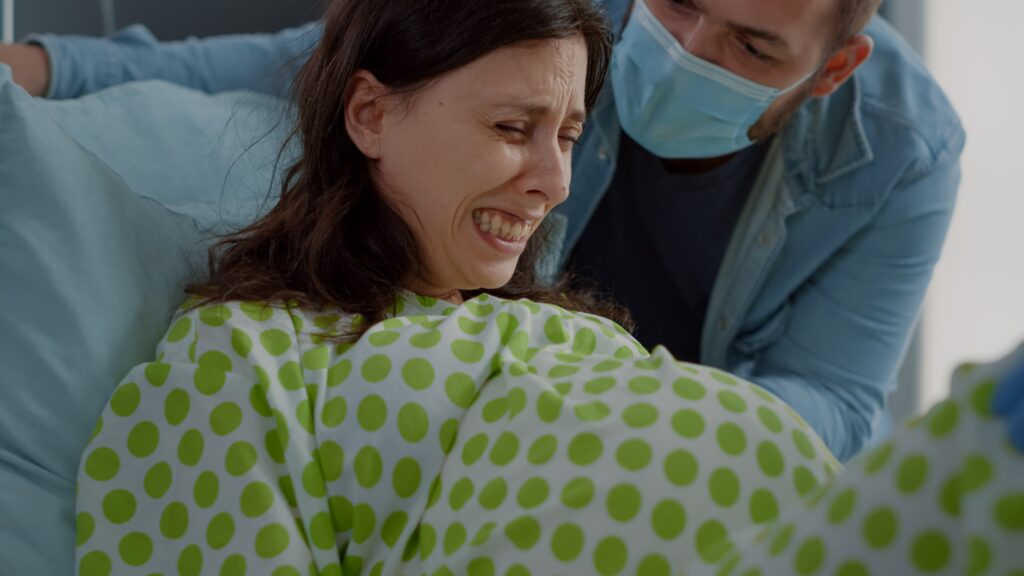 Una mujer con una bata de hospital verde con puntos blancos está acostada en una cama, con el rostro contraído por el dolor, aparentemente durante el parto. Un hombre con mascarilla azul la apoya desde detrás.