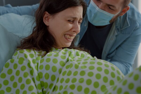 Una mujer con una bata de hospital verde con puntos blancos está acostada en una cama, con el rostro contraído por el dolor, aparentemente durante el parto. Un hombre con mascarilla azul la apoya desde detrás.