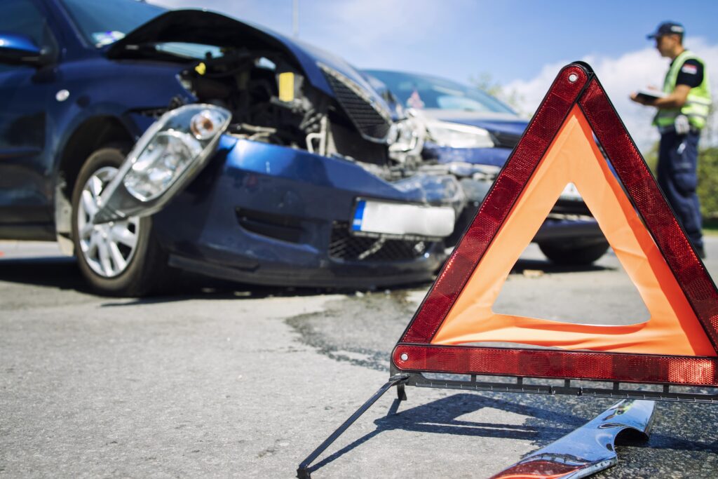 En primer plano, un triángulo de advertencia rojo y naranja en la carretera. Al fondo, se ven dos coches azules que han sufrido daños frontales por un accidente, y una persona con uniforme de policía o de tráfico está de pie cerca de ellos tomando notas.