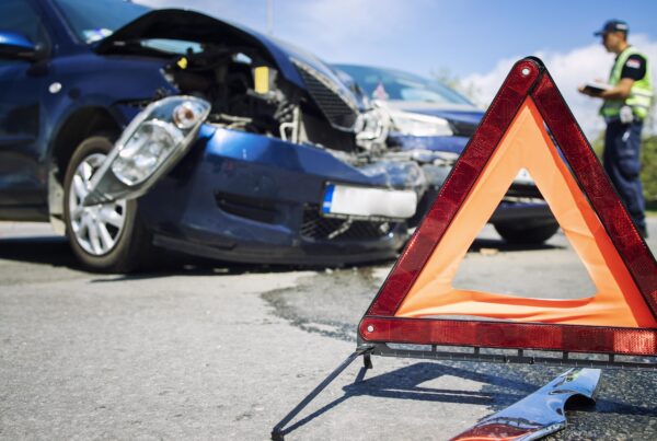 En primer plano, un triángulo de advertencia rojo y naranja en la carretera. Al fondo, se ven dos coches azules que han sufrido daños frontales por un accidente, y una persona con uniforme de policía o de tráfico está de pie cerca de ellos tomando notas.