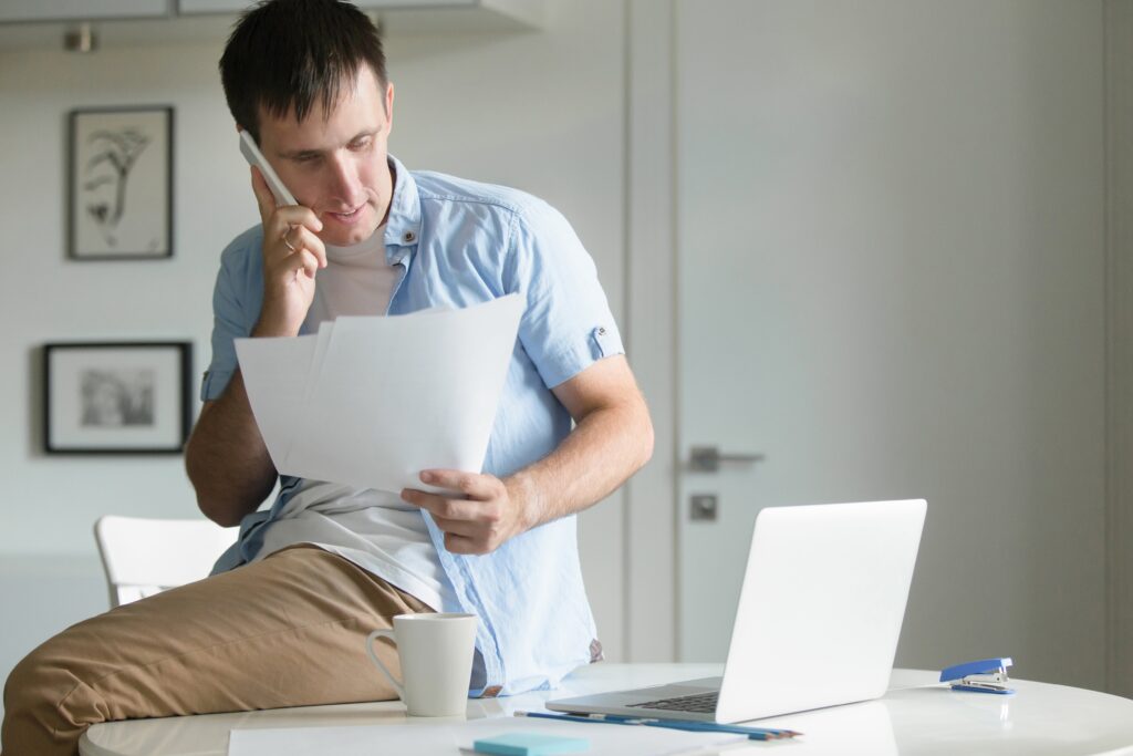 Un hombre joven, vestido con una camisa azul claro y pantalones beige, está sentado en el borde de un escritorio blanco, hablando por teléfono mientras revisa unos documentos. Hay un portátil, una taza y otros objetos de oficina sobre la mesa.