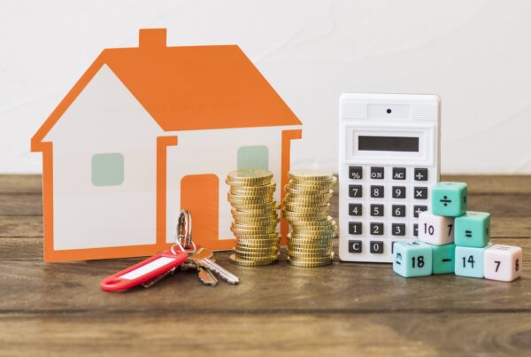 Una maqueta de casa de color naranja y blanco, junto a dos pilas de monedas doradas y un juego de llaves en primer plano. Detrás, hay una calculadora blanca y varios dados de colores con números, lo que sugiere conceptos de inversión inmobiliaria, ahorro o hipotecas.