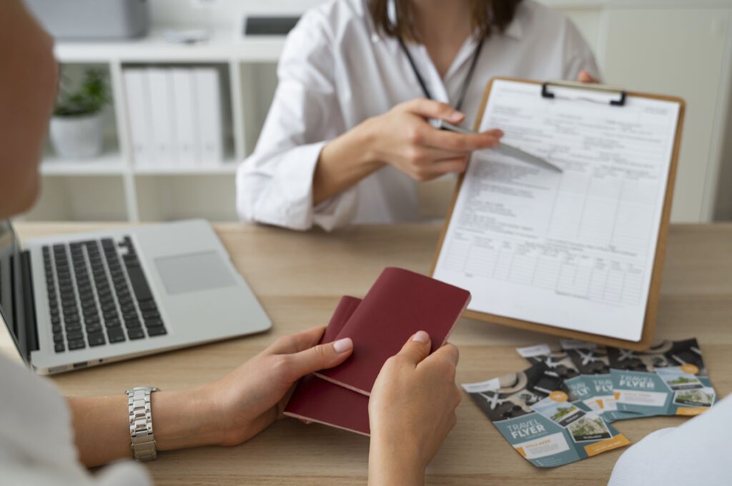 Dos personas sentadas a un escritorio. Una de ellas, con una camisa blanca, señala un documento en un portapapeles mientras la otra persona sostiene dos pasaportes rojos. En la mesa también hay un portátil y folletos de "Travel Flyer", indicando una gestión de viajes o visados.