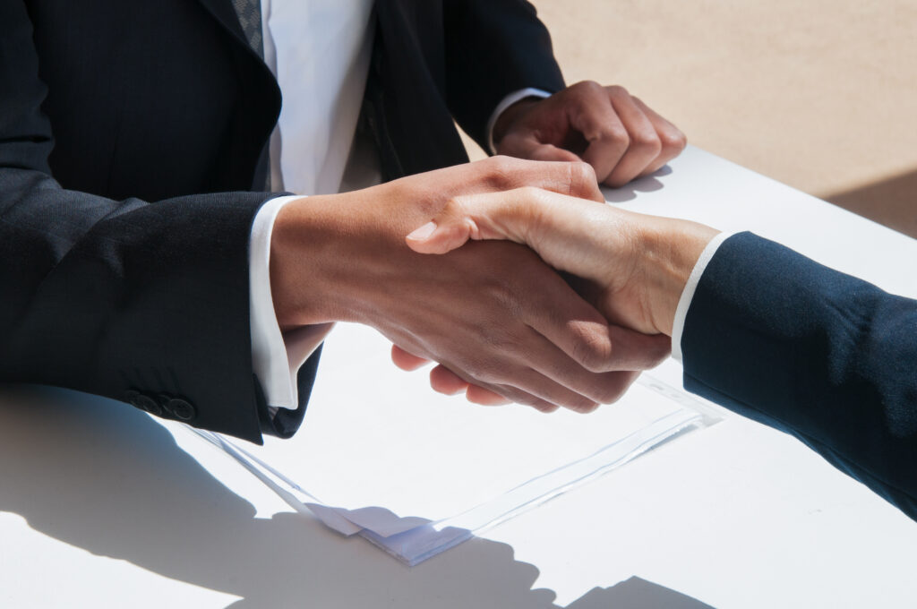 Dos personas vestidas formalmente se dan la mano sobre una mesa blanca. La imagen se centra en el apretón de manos, que sugiere un acuerdo o saludo de negocios.