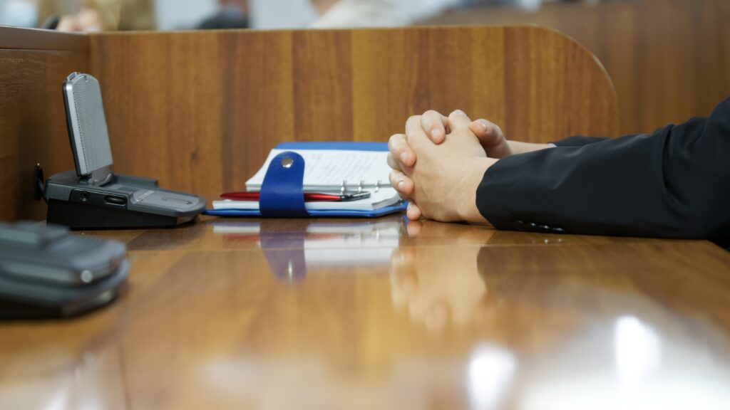 Unas manos entrelazadas sobre una mesa de madera brillante, en un entorno de oficina. Hay una agenda azul con un bolígrafo rojo y un dispositivo electrónico en la mesa, lo que sugiere una reunión o una espera tensa. El fondo es un mostrador de madera.