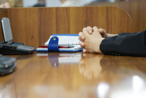 Unas manos entrelazadas sobre una mesa de madera brillante, en un entorno de oficina. Hay una agenda azul con un bolígrafo rojo y un dispositivo electrónico en la mesa, lo que sugiere una reunión o una espera tensa. El fondo es un mostrador de madera.