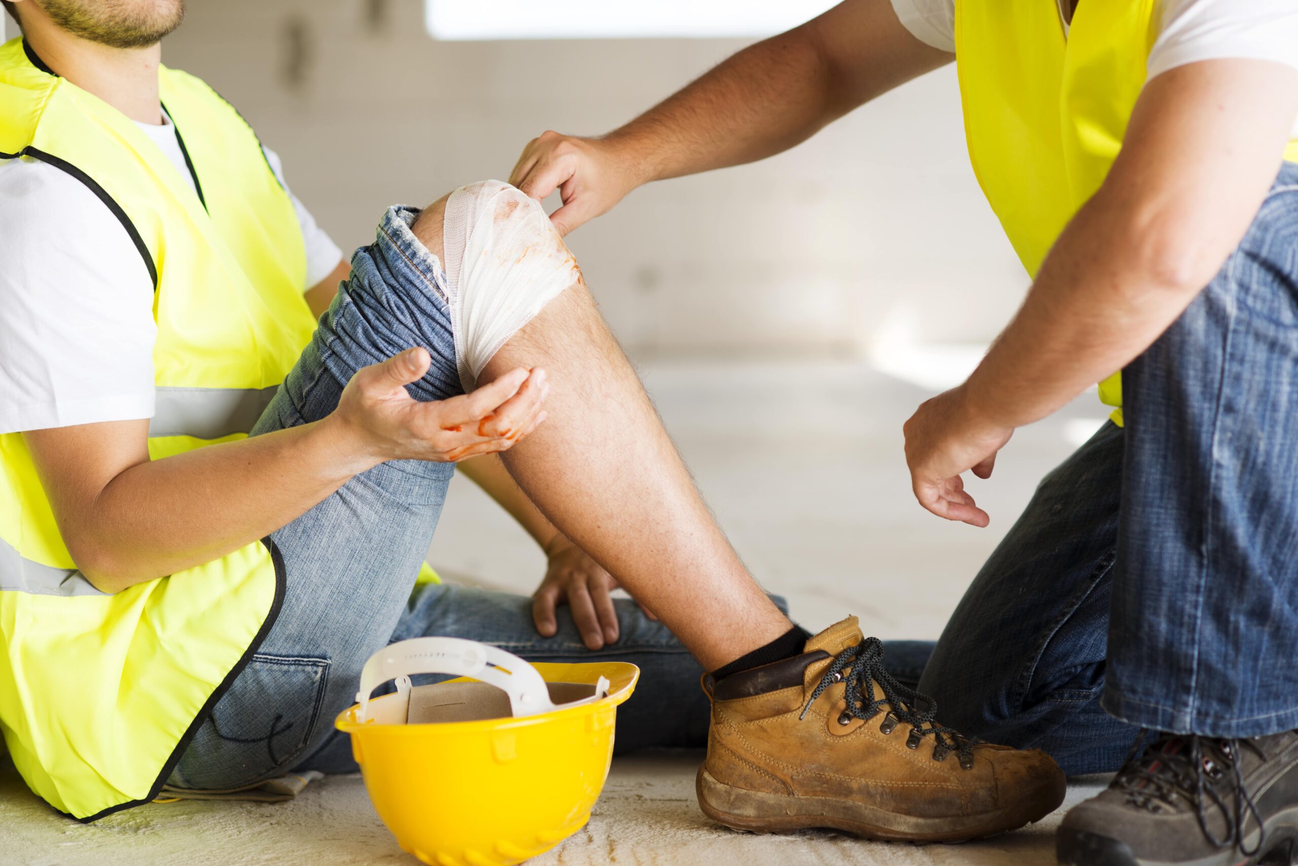 Un trabajador de la construcción con un chaleco reflectante amarillo sentado en el suelo, con una venda visible en su rodilla. Otro trabajador, también con chaleco reflectante, está agachado a su lado ayudándolo. Un casco amarillo de construcción y unas botas de trabajo se ven en el suelo. La imagen representa un accidente laboral.