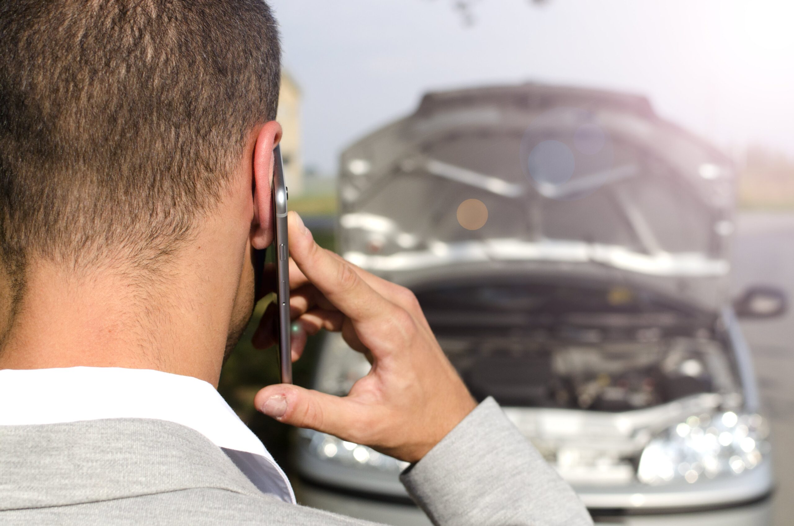La parte trasera de la cabeza de un hombre hablando por un teléfono móvil, con un coche plateado con el capó abierto y vapor saliendo, desenfocado en el fondo. La imagen sugiere que el hombre está lidiando con una avería o accidente de coche.