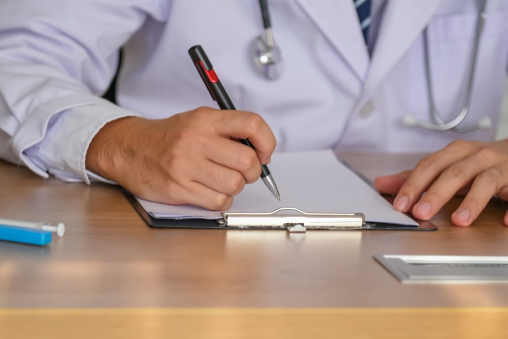 Un médico con bata blanca y estetoscopio, sentado en un escritorio de madera, escribiendo en un portapapeles con un bolígrafo negro y rojo. Sus manos son el foco principal, lo que sugiere el registro de información médica.