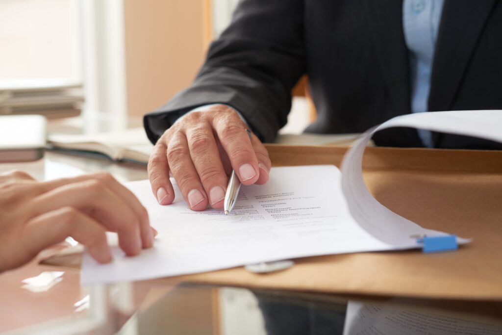 Dos personas están sentadas a una mesa. Una de ellas, con traje oscuro, sostiene un bolígrafo y señala un documento que está siendo revisado por la otra persona. La escena sugiere la firma o revisión de un contrato o documento legal.