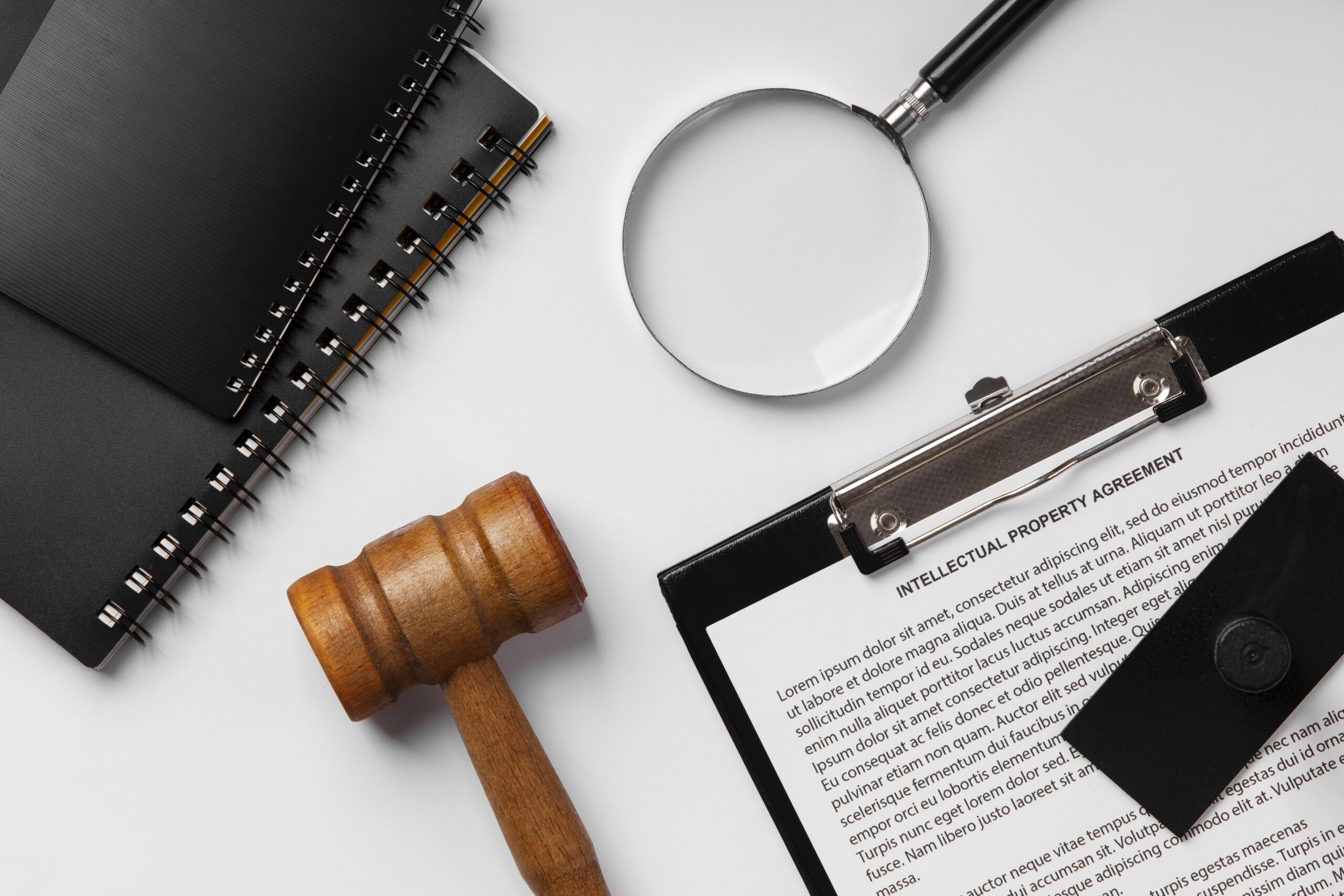 Una vista cenital de objetos relacionados con el ámbito legal sobre una superficie blanca. Se aprecia un mazo de madera de juez, una lupa, una pila de cuadernos negros de espiral y un portapapeles con un documento titulado "INTELLECTUAL PROPERTY AGREEMENT".